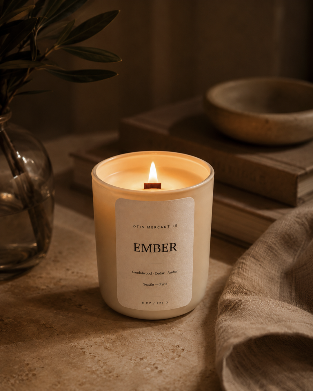Lit candle labeled 'Ember' on a surface with a warm glow, surrounded by a plant and wooden bowl.