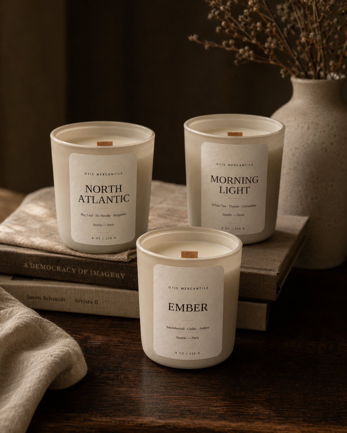 Three candles labeled 'North Atlantic', 'Morning Light', and 'Ember' on a wooden surface with books and a vase in the background.
